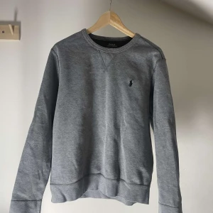 Grå sweatshirt från Polo Ralph Lauren - Säljer en stilren grå sweatshirt från Polo Ralph Lauren med deras ikoniska logga broderad på bröstet. Tröjan har långa ärmar och en klassisk rund halsringning. Perfekt för en avslappnad och snygg look.