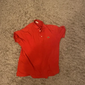Röd pikétröja från Lacoste - Säljer en klassisk röd pikétröja från Lacoste med den ikoniska krokodilloggan på bröstet. Tröjan har korta ärmar och en traditionell krage med knappar. Perfekt för en stilren och avslappnad look.den sitter som en large