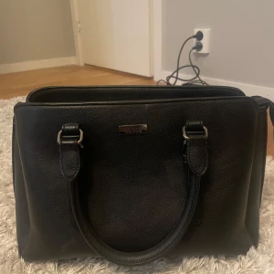 Svart handväska från Don Donna - Snygg svart handväska från Don Donna i skinn. Väskan har två robusta handtag och en stilren design med en metallplatta med logotyp på framsidan. Perfekt för att bära dina dagliga nödvändigheter med stil.