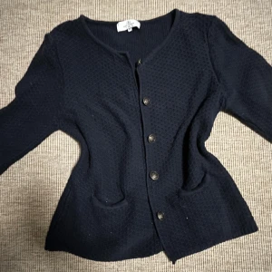 Svart kofta från Flash Woman - Säljer en elegant svart kofta från Flash Woman. Koftan har en fin struktur och knäpps med knappar framtill. Den har långa ärmar och en klassisk rund halsringning. Perfekt för en stilren look.