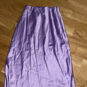 Lila sidenkjol - Elegant lila långkjol i sidenmaterial. Perfekt för en stilren look med sin släta och glänsande yta. Kjolen har en enkel design utan extra detaljer, vilket gör den lätt att matcha med olika toppar.