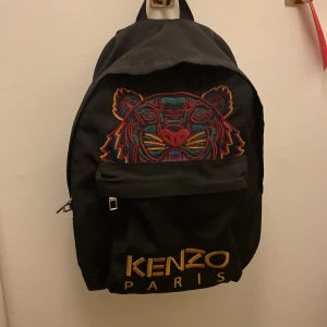 Svart ryggsäck från Kenzo - Snygg svart ryggsäck från Kenzo med ett coolt tigerbroderi i färgglada nyanser på framsidan. Väskan har en praktisk dragkedja och justerbara axelremmar. Perfekt för att bära dina grejer med stil.