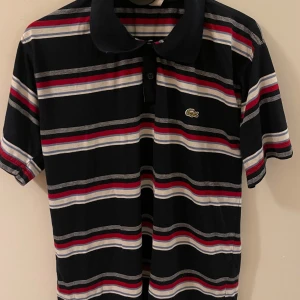 Randig pikétröja från Lacoste - Snygg randig pikétröja från Lacoste i svart, röd, vit och grå. Tröjan har korta ärmar och en klassisk krage med knappar. Perfekt för en stilren och avslappnad look.