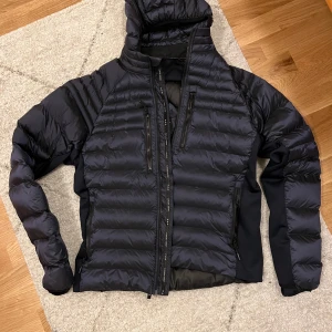 Svart dunjacka från Canada Goose - Säljer en svart dunjacka från Canada Goose med huva och flera praktiska dragkedjor. Jackan har en snygg quiltad design och ett märke på ärmen. Perfekt för kyliga dagar.