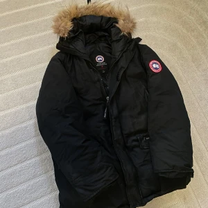 Svart parkajacka från Canada Goose expedition  - Säljer en svart parkajacka från Canada Goose med pälskantad huva. Jackan har en dragkedja och är perfekt för kalla vinterdagar. Den har en klassisk design med en logotyp på ärmen.