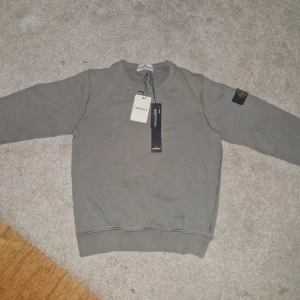Grå sweatshirt från Stone Island - Säljer en stilren grå sweatshirt från Stone Island. Tröjan har en rund halsringning och ett ikoniskt märke på ärmen. Perfekt för en avslappnad look.