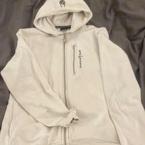 Vit hoodie från Sail Racing - Säljer en vit hoodie från Sail Racing med dragkedja och huva. Den har en bröstficka med dragkedja och logotyp på bröstet. Perfekt för en avslappnad stil.