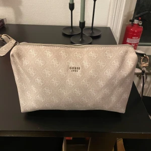 Beige axelväska från Guess - Snygg beige axelväska från Guess med ett diskret mönster och logotyp framtill. Väskan har en justerbar axelrem och dragkedja upptill. Perfekt för att bära dina nödvändigheter med stil.