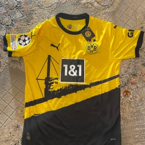 Gul Borussia Dortmund fotbollströja - Säljer en gul Borussia Dortmund fotbollströja från Puma med svart tryck och detaljer. Tröjan har korta ärmar och klubbmärket BVB 09 på bröstet. Nummer 11 och namnet Reus är tryckt på baksidan. Perfekt för fans av laget!
