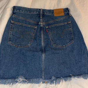 Jeanskjol från Levi's - Säljer en snygg jeanskjol från Levi's i klassisk blå denim. Kjolen har en rå kant nertill och femficksdesign med knapp och dragkedja framtill. Perfekt för en avslappnad stil.