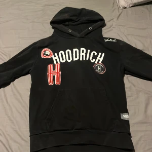 Svart hoodie från Hoodrich - Säljer en svart hoodie från Hoodrich med vita och röda detaljer. Hoodien har en stor ficka framtill och texten 'HOODRICH' i vitt på bröstet. Perfekt för en avslappnad stil.