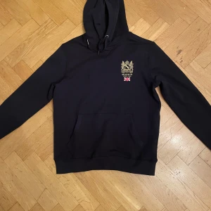 marinblå hoodie från Morris - Snygg marinblå hoodie från Morris med broderad logga och brittisk flagga på bröstet. Perfekt för en stilren och avslappnad look. Den har en klassisk känguruficka och justerbar huva. använd max två gånger. Skriv för frågor eller funderingar 