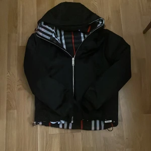 Svart jacka från Burberry - Säljer en snygg svart jacka från Burberry med dragkedja och huva. Jackan har ett klassiskt rutigt mönster på insidan.tar byten .har ett mönster som man ser på sista bilden 