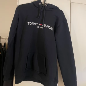 Svart hoodie från Tommy Hilfiger - Säljer en stilren svart hoodie från Tommy Hilfiger med logotyp och text 'Est. 1985' på bröstet. Tröjan har en klassisk känguruficka och justerbar huva med snören. Perfekt för en avslappnad look.