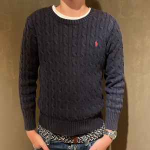 Mörkblå stickad tröja från Polo Ralph Lauren - Säljer en snygg mörkblå stickad tröja från Polo Ralph Lauren med kabelstickat mönster och röd logga på bröstet. Tröjan har långa ärmar och ribbade muddar vid ärmslut och nederkant. Perfekt för en stilren look!