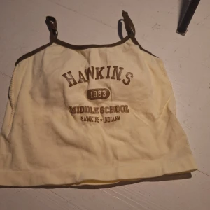 Gul linne med tryck - Säljer en gul linne med brunt tryck 'Hawkins Middle School 1983'. Den har smala axelband och bruna kanter. Perfekt för en avslappnad stil. Den har även inbyggda inlägg.