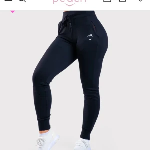 Svarta mjukisbyxor från Peach - Snygga svarta mjukisbyxor från Peach i en skön blandning av nylon och elastan. Perfekta för en aktiv livsstil med en tight passform och elastiska muddar vid anklarna. Diskret logga på sidan ger en stilren look.