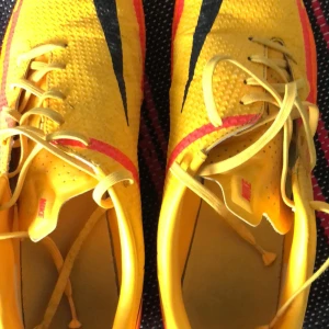 Gula Nike Phantom fotbollsskor - Snygga gula Nike Phantom fotbollsskor med svarta och röda detaljer. Skorna har snörning och är designade för optimal prestanda på planen.