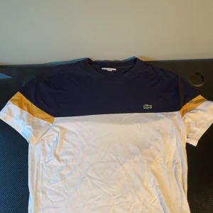  Vit och blå Lacoste T-shirt - Säljer en snygg t-shirt från Lacoste i blått och vitt med gula detaljer på ärmarna. T-shirten har en normal passform och korta ärmar. Perfekt för en avslappnad stil.