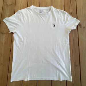 Vit t-shirt från Ralph Lauren - Säljer en klassisk vit t-shirt från Ralph Lauren med det ikoniska broderade logotypen på bröstet. T-shirten är kortärmad och tillverkad i mjuk bomull för en bekväm passform.