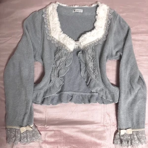 Axes framme Grå kofta med spets och rosett - Charmig grå kofta från Axes Femme med spetsdetaljer och volanger. Den har en fluffig krage och söta rosetter med pärlor vid ärmsluten. Perfekt för en romantisk look.