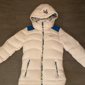 Vit dunjacka från Craft - Säljer en snygg vit dunjacka från Craft med blå och gula detaljer. Jackan har en cool mönstrad insida och en broderad häst på huvan. Perfekt för kalla dagar med sin långa ärmar och dragkedja framtill. Håll dig varm och stilren! 🐴Jackan är pefekt för skidåkning och vardagsliv och är nästan oanvänd