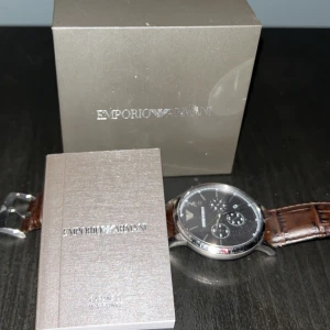 Emporio Armani herrklocka med svart läderrem - Stilren herrklocka från Emporio Armani med brun läderrem och silverfärgad urtavla. Klockan har flera små urtavlor och en elegant design som passar perfekt för den modemedvetne. Levereras med originalförpackning och manual.
