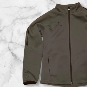 Grön jacka från Hummel - Säljer en grön jacka från Hummel med dragkedja framtill. Jackan har långa ärmar och en stilren design med diskreta logodetaljer. Perfekt för en sportig look.