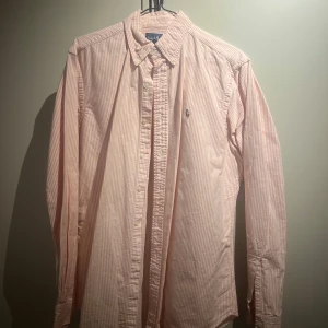 Rosa randig skjorta från Ralph Lauren - Säljer en klassisk rosa och vit randig skjorta från Ralph Lauren. Skjortan har långa ärmar och en broderad logotyp på bröstet. Perfekt för en stilren look.