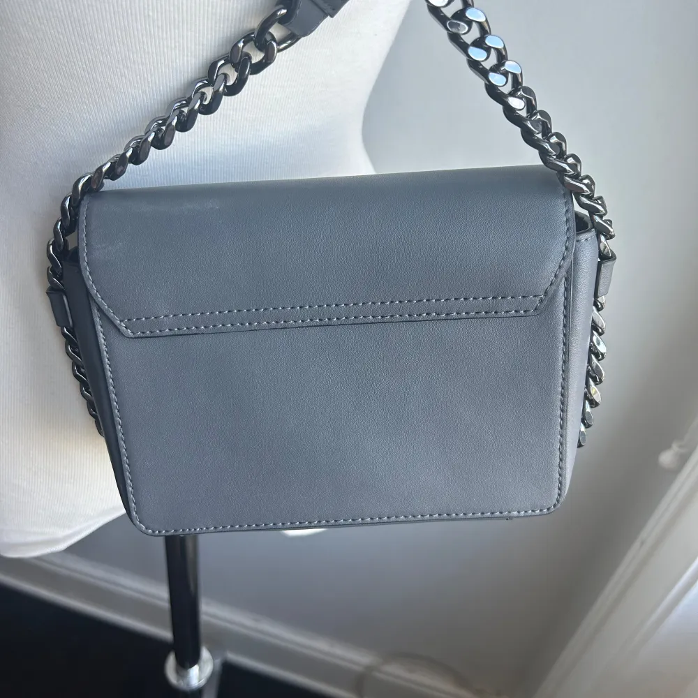 Säljer en Karl Lagerfeld stilren grå axelväska med en cool kedjedetalj på axelremmen. Väskan har en elegant design med ett metallmärke på framsidan. Perfekt för både vardag och fest!. Laukut & Käsilaukut.