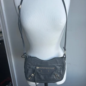 Balenciaga väska - Säljer min Balenciaga ”Metallic Edge Hip Bag”. Väskan har en dragkedja framtill och små spännen i guld. Perfekt för både vardag och fest! Den är i bra skick och har en justerbar axelrem.