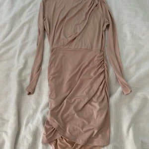 Beige klänning från Gina Tricot - Säljer en snygg beige/rosa klänning från Gina Tricot i storlek S. Klänningen har en asymmetrisk design med långärmade ärmar och en draperad detalj på sidan. 