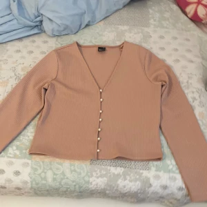 Beige ribbad kofta med pärlknappar - Säljer en superfin beige kofta med ribbad struktur och v-ringning. Den har långa ärmar och är dekorerad med eleganta pärlknappar framtill. Perfekt för både vardag och fest! 🧡