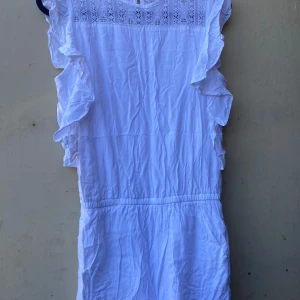 Vit klänning med spetsdetaljer - Säljer en superfin vit byxdress med spetsdetaljer upptill och volanger längs ärmarna. Byxdressen är ärmlös och har en elastisk midja för en bekväm passform. Perfekt för sommarens alla fester eller en dag på stranden! 🌸