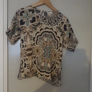 Mönstrad blus i beige och blå nyanser - Säljer en superfin blus med ett unikt mönster i beige, blå och bruna nyanser. Blusen har korta ärmar och en lös passform, perfekt för både vardag och fest.