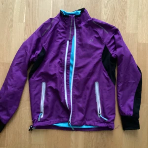 Lila och svart vindjacka från Pro Touch - Säljer en snygg lila vindjacka från Pro Touch med svarta detaljer. Jackan har en dragkedja framtill och två praktiska fickor med dragkedjor. Insidan är i en fräsch turkos färg som ger en cool kontrast. Perfekt för blåsiga dagar och utomhusaktiviteter!