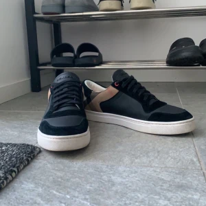 Burberry skor - Säljer ett par snygga svarta sneakers från Burberry i skick 8/10. De har en klassisk rutig detalj på hälen och är gjorda i skinn och mocka. Perfekta för både vardag och fest! Skriv för bilder, frågor mm!