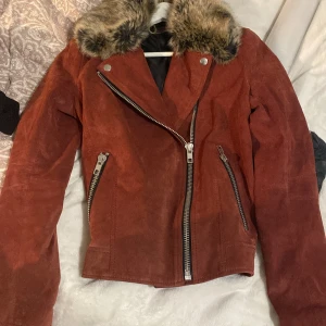 Rostfärgad mockajacka från Jofama - Säljer en snygg rostfärgad mockajacka från Jofama med en lyxig fuskpäls på kragen. Jackan har en cool asymmetrisk dragkedja och två fickor med dragkedjor. Perfekt för höst och vår när du vill ha en stilren look. Passar både till vardags och fest!