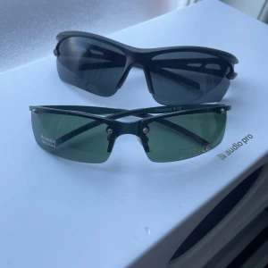 Solglasögon i sportig stil - Två par solglasögon i sportig stil. Ett par med svarta bågar och mörka linser, perfekt för träning eller sommaraktiviteter. Det andra paret har gröna linser och är behandlade mot nickel, vilket gör dem skonsamma mot huden. Båda paren har en modern och funktionell design.