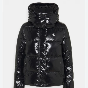 Svart glansig pufferjacka från Calvin Klein - Snygg svart pufferjacka från Calvin Klein. Jackan har en hög krage och dragkedja framtill. Perfekt för kyliga dagar och ger en stilren look. Den har en liten CK-logga framtill.