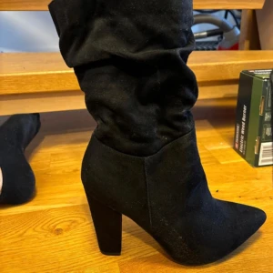 Svarta stövletter med klack - Snygga svarta stövletter i mocka med hög klack och spetsig tå. Perfekta för höst och vinter, dessa boots har en stilren design som passar både till vardags och fest. Tillverkade i Kina och i mycket bra skick.