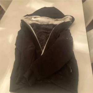 Svart jacka från Moose Knuckles - Säljer en svart jacka från Moose Knuckles i mycket bra skick. Den har en mysig vit foder och en dragkedja framtill. Jackan är perfekt för kyligare dagar och har snygga detaljer som märkesloggan på ärmen. Passar både till vardags och lite finare tillfällen.Jackan är i St M men passar S