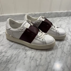 Valentino Garavani sneakers i vitt och vinrött - Säljer ett par snygga Valentino Garavani sneakers i vitt skinn med vinröda detaljer. Skorna har coola nitar runt hälen och en bred rem över snörningen. Använda men i bra skick, har tyvärr inte kvitto eller og saker för att min mamma hade inte sparat det men hör av dig vid intresse samt frågor, pris är ej hugget i sten😉🤩