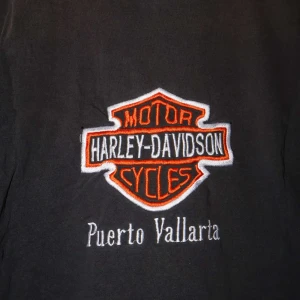 Svart Harley-Davidson t-shirt från Yazbek - Säljer en svart t-shirt från Yazbek med det klassiska Harley-Davidson-logotypen broderad på bröstet och ett större bak på ryggen. T-shirten är i bomull och har korta ärmar, perfekt för en avslappnad stil.