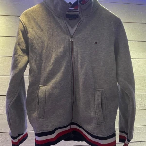 Grå zip-up tröja från Tommy Hilfiger - Säljer en snygg grå zip-up tröja från Tommy Hilfiger. Den har långa ärmar och ribbade muddar med röd, vit och blå randig detalj. Perfekt för en casual look eller en kylig dag. Tröjan har en liten logga på bröstet och en större på ärmen. Passar både till vardags och lite finare tillfällen.