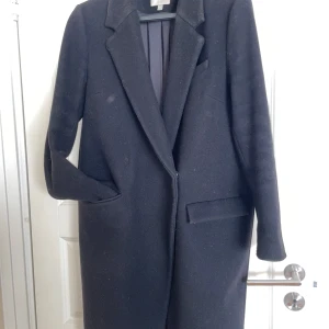 Svart kappa från dagmar - Säljer en elegant svart kappa från Dagmar i storlek M. Den är tillverkad av 70% ull och 30% polyester, vilket gör den perfekt för höst och vinter. Kappan har en klassisk design med långa ärmar och en stilren passform. Perfekt för både vardag och festliga tillfällen!