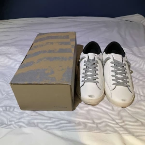 Vita sneakers från Golden Goose - Säljer ett par snygga vita sneakers från Golden Goose i nytt skick. De har grå snörning och en cool stjärndetalj på sidan. Skorna kommer med originalkartong och påse. 