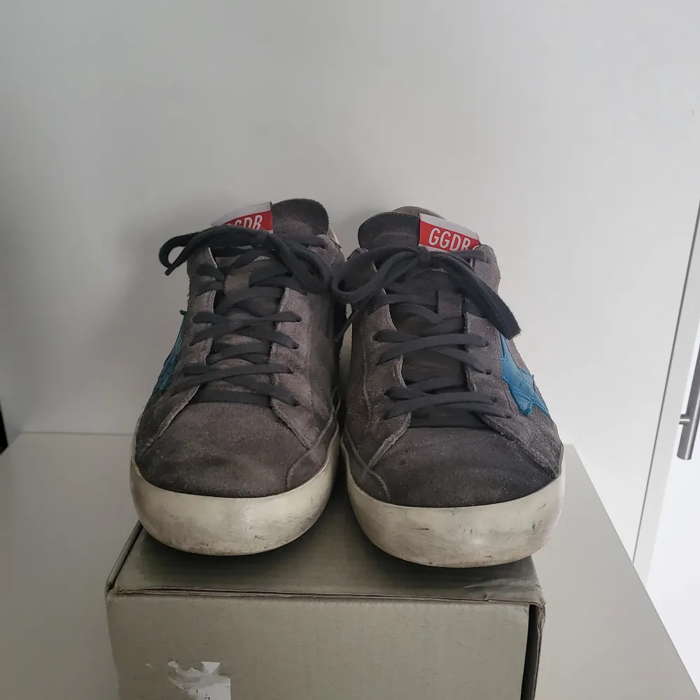 Säljer ett par grå sneakers från Golden Goose i bra skick. De har en cool blå stjärna på sidan och vit häl med märkesnamnet. Skorna har snörning och är gjorda i mocka. Perfekta för en avslappnad stil med en touch av lyx. Kommer med originalförpackning.. Skor.