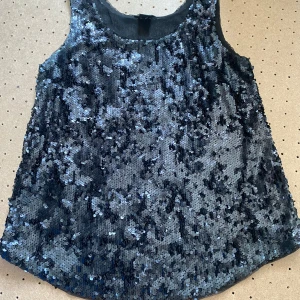 Svart paljettopp från Lindex - Snygg svart paljettopp från Lindex i storlek XS. Perfekt för festliga tillfällen med sitt glittrande utseende. Toppen är ärmlös och har en lös passform som ger en bekväm känsla. Passar perfekt till både kjol och byxor för en glamorös look! Bra till jul, nyår och andra fester.