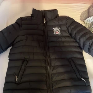Svart pufferjacka - Säljer en svart pufferjacka från Lonsdale i mycket bra skick. Jackan har en dragkedja framtill och ett broderat emblem på bröstet. Perfekt för kyliga dagar med sin vadderade design. Den har långa ärmar och två fickor med dragkedja.
